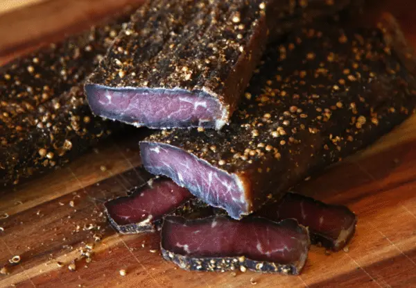 Close-up of traditional South African biltong sliced on a wooden cutting board, seasoned with coriander and black pepper. Dark, cured meat texture with visible spice crust.